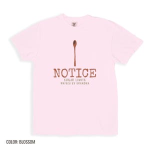 Blossom Comfort Colors t-shirt printed with the words Notice Sugar Limits Waived By Grandma and a drawing of a painted wooden spoon crossed over a cinnamon stick bundle, folded flat lay