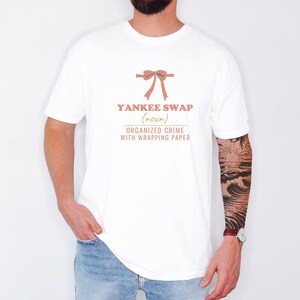 White Comfort Colors t-shirt printed with the words Yankee Swap (Noun) Organized Crime With Wrapping Paper and a drawing of a large red comical bow with long satin ribbon tails, worn by a man