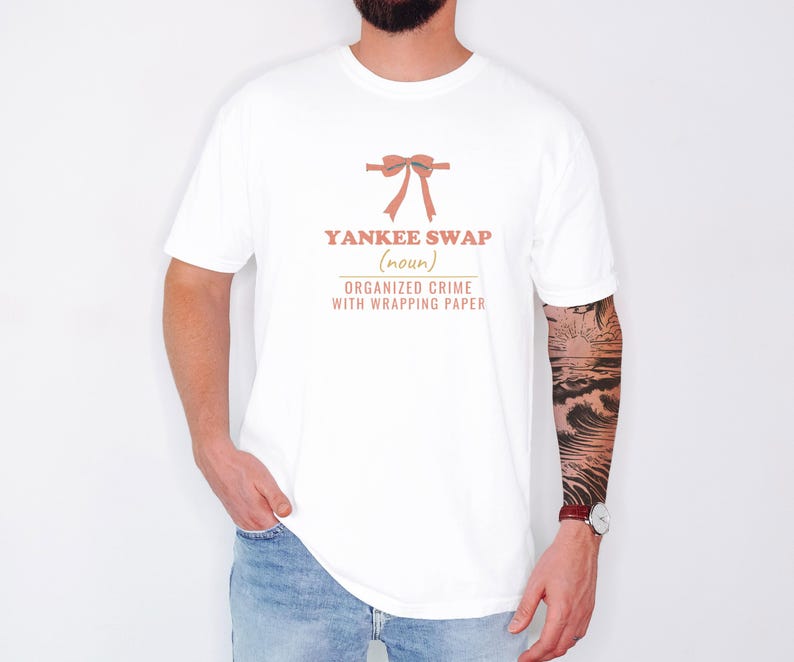 White Comfort Colors t-shirt printed with the words Yankee Swap (Noun) Organized Crime With Wrapping Paper and a drawing of a large red comical bow with long satin ribbon tails, worn by a man
