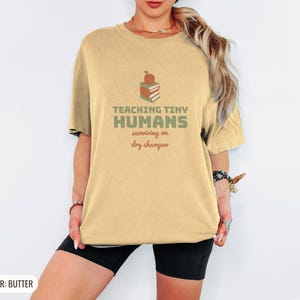 Butter Comfort Colors t-shirt printed with the words Teaching Tiny Humans Surviving On Dry Shampoo and a drawing of a small neat stack of three closed hardcover books with PLAIN blank solid-colour spines an, worn by a woman