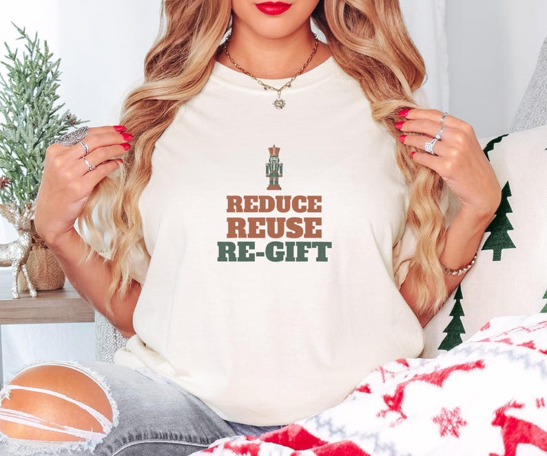 Ivory Comfort Colors t-shirt printed with the words Reduce Reuse Re-Gift and a drawing of a vintage wooden nutcracker soldier standing beside a folded green paper gift tag, worn by a woman