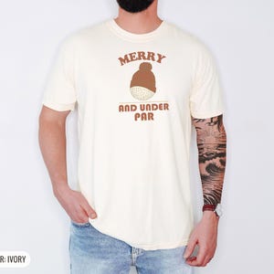 Ivory Comfort Colors t-shirt printed with the words Merry And Under Par and a drawing of a dimpled golf ball wearing a tiny red and white striped knit winter beanie with a green p, worn by a man