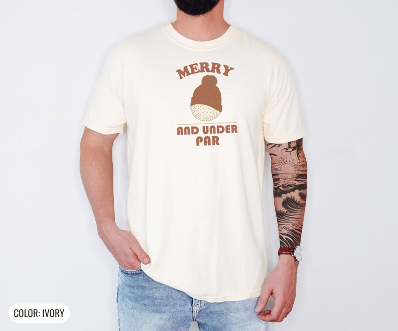 Ivory Comfort Colors t-shirt printed with the words Merry And Under Par and a drawing of a dimpled golf ball wearing a tiny red and white striped knit winter beanie with a green p, worn by a man