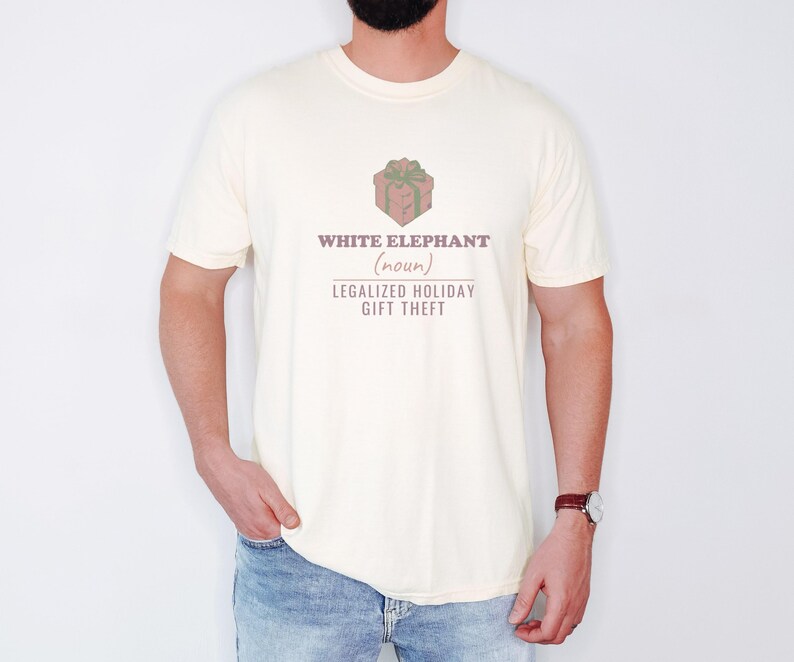 Ivory Comfort Colors t-shirt printed with the words White Elephant (Noun) Legalized Holiday Gift Theft and a drawing of a shiny wrapped gift parcel with a giant comical polka-dot bow, worn by a man