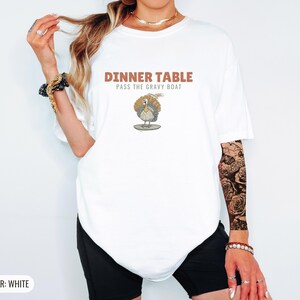 White Comfort Colors t-shirt printed with the words Dinner Table Pass The Gravy Boat and a drawing of a golden roast turkey on a platter, worn by a woman