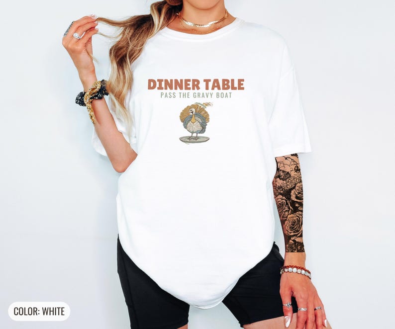 White Comfort Colors t-shirt printed with the words Dinner Table Pass The Gravy Boat and a drawing of a golden roast turkey on a platter, worn by a woman