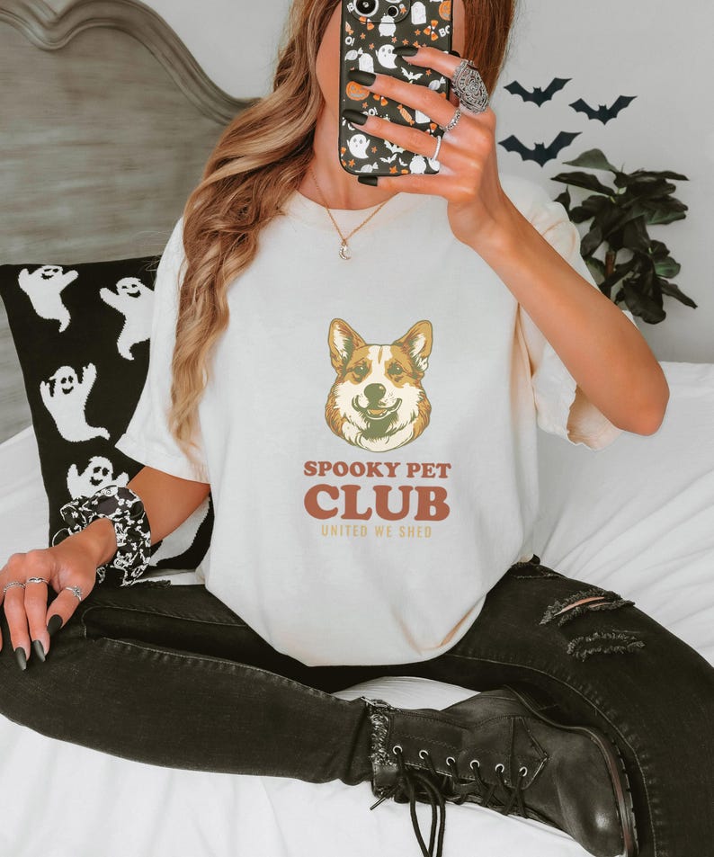 Ivory Comfort Colors t-shirt printed with the words Spooky Pet Club United We Shed and a drawing of a circular crest showing a corgi spirit and a black feline nose-to-nose, worn by a woman