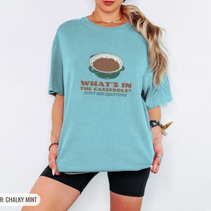 Chalky Mint Comfort Colors t-shirt printed with the words What'S In The Casserole? Don'T Ask Questions and a drawing of a bubbly baked casserole in a rectangular ceramic baking dish topped with crispy fried oni, worn by a woman