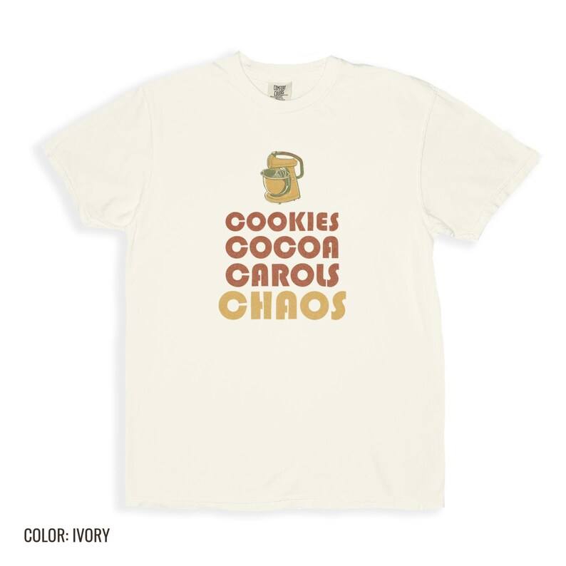 Ivory Comfort Colors t-shirt printed with the words Cookies Cocoa Carols Chaos and a drawing of a retro metal hand mixer with steel beaters, folded flat lay