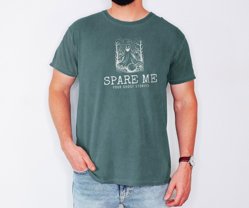 Blue Spruce Comfort Colors t-shirt printed with the words Spare Me Your Ghost Stories and a drawing of a friendly sheet ghost rolling a bowling ball towards a set of bowling pins, worn by a man