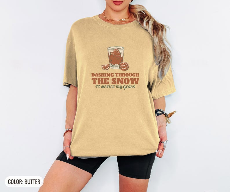 Butter Comfort Colors t-shirt printed with the words Dashing Through The Snow To Refill My Glass and a drawing of a heavy crystal whiskey tumbler with two floating ice cubes beside a sprig of pine needles, worn by a woman