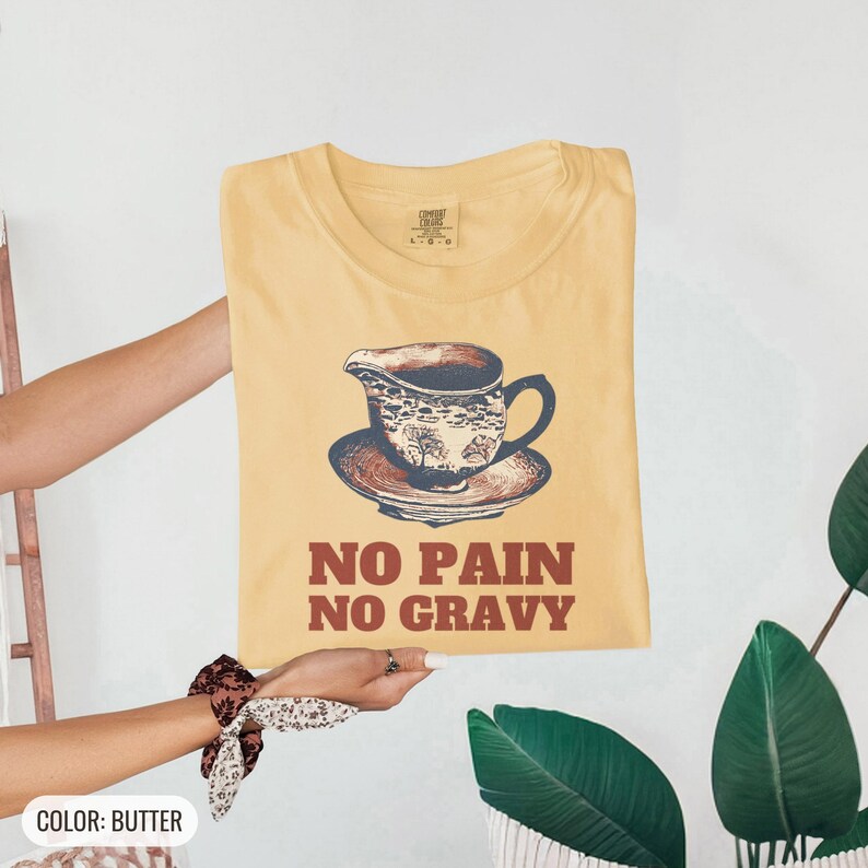 Butter Comfort Colors t-shirt printed with the words No Pain No Gravy and a drawing of a ceramic gravy boat with a small puddle of gravy underneath, folded flat lay
