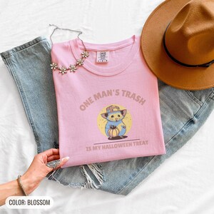 Blossom Comfort Colors t-shirt printed with the words One Man'S Trash Is My Halloween Treat and a drawing of a scruffy opossum wearing a tiny plastic orange pumpkin trick-or-treat bucket on its head, folded flat lay