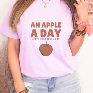 Orchid Comfort Colors t-shirt printed with the words An Apple A Day Keeps The Chaos Away and a drawing of a shiny red apple with a single green leaf and a tiny worm peeking out, worn by a woman