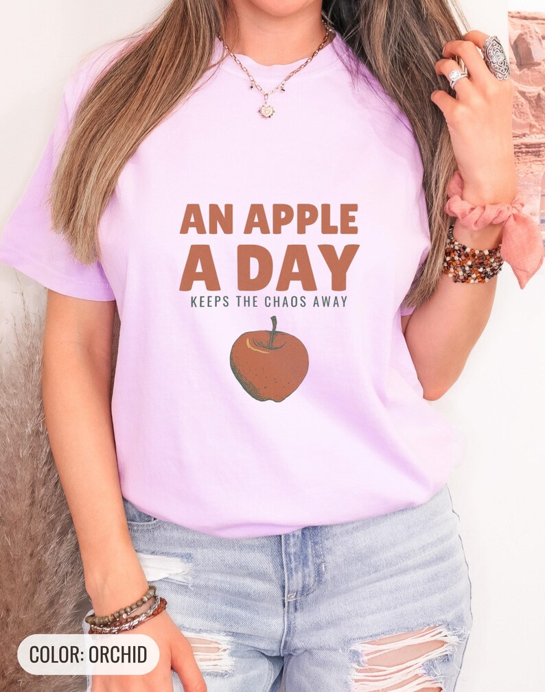 Orchid Comfort Colors t-shirt printed with the words An Apple A Day Keeps The Chaos Away and a drawing of a shiny red apple with a single green leaf and a tiny worm peeking out, worn by a woman
