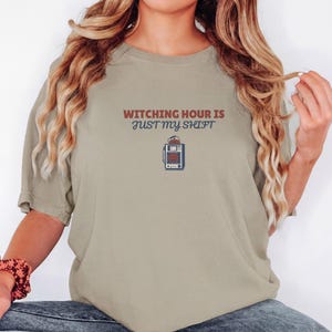 Khaki Comfort Colors t-shirt printed with the words Witching Hour Is Just My Shift and a drawing of a retro medical defibrillator machine decorated with a spooky jack-o-lantern face on its m, worn by a woman