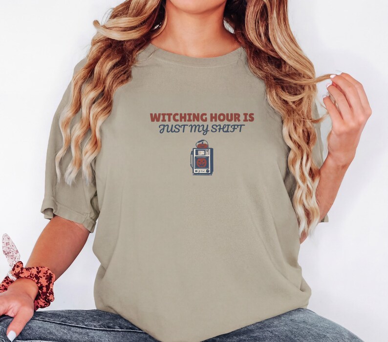 Khaki Comfort Colors t-shirt printed with the words Witching Hour Is Just My Shift and a drawing of a retro medical defibrillator machine decorated with a spooky jack-o-lantern face on its m, worn by a woman