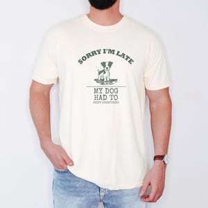 Ivory Comfort Colors t-shirt printed with the words Sorry I'M Late My Dog Had To Sniff Everything and a drawing of a brown and white terrier standing in a puddle, worn by a man