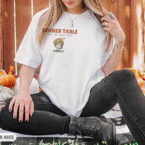 White Comfort Colors t-shirt printed with the words Dinner Table Pass The Gravy Boat and a drawing of a golden roast turkey on a platter, worn by a woman