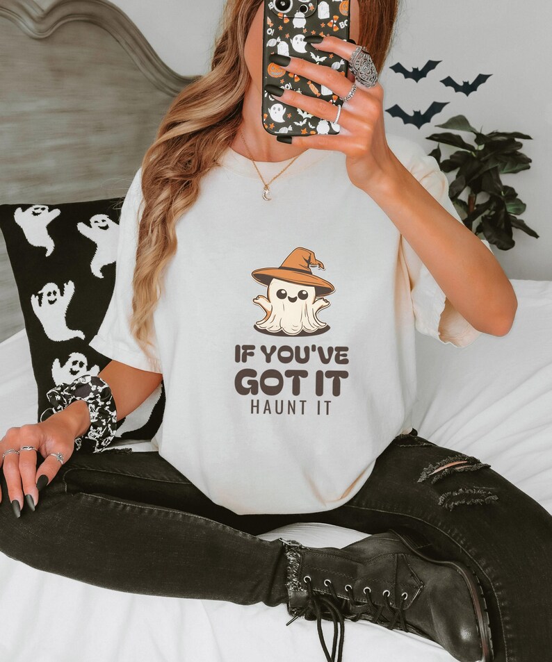 Ivory Comfort Colors t-shirt printed with the words If You'Ve Got It Haunt It and a drawing of a cute retro cartoon ghost wearing a witch hat, worn by a woman