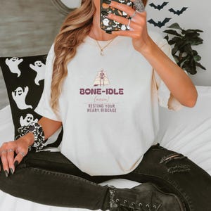 Ivory Comfort Colors t-shirt printed with the words Bone-Idle (Noun) Resting Your Weary Ribcage and a drawing of a skeleton completely tucked under a thick, fluffy duvet in a wooden four-poster bed, worn by a woman