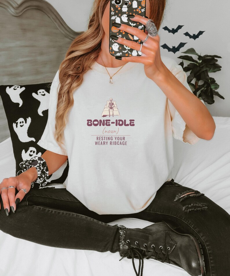 Ivory Comfort Colors t-shirt printed with the words Bone-Idle (Noun) Resting Your Weary Ribcage and a drawing of a skeleton completely tucked under a thick, fluffy duvet in a wooden four-poster bed, worn by a woman