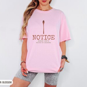 Blossom Comfort Colors t-shirt printed with the words Notice Sugar Limits Waived By Grandma and a drawing of a painted wooden spoon crossed over a cinnamon stick bundle, worn by a woman