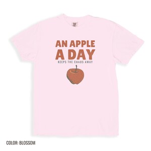 Blossom Comfort Colors t-shirt printed with the words An Apple A Day Keeps The Chaos Away and a drawing of a shiny red apple with a single green leaf and a tiny worm peeking out, folded flat lay
