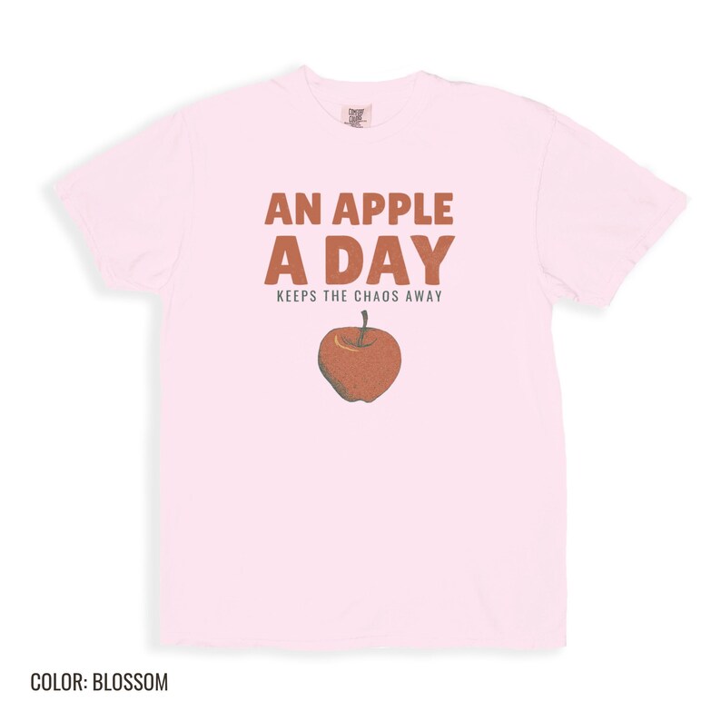Blossom Comfort Colors t-shirt printed with the words An Apple A Day Keeps The Chaos Away and a drawing of a shiny red apple with a single green leaf and a tiny worm peeking out, folded flat lay