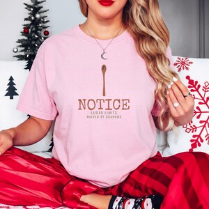 Blossom Comfort Colors t-shirt printed with the words Notice Sugar Limits Waived By Grandma and a drawing of a painted wooden spoon crossed over a cinnamon stick bundle, worn by a woman