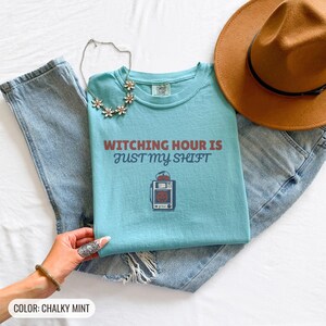 Chalky Mint Comfort Colors t-shirt printed with the words Witching Hour Is Just My Shift and a drawing of a retro medical defibrillator machine decorated with a spooky jack-o-lantern face on its m, folded flat lay
