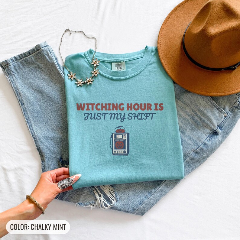 Chalky Mint Comfort Colors t-shirt printed with the words Witching Hour Is Just My Shift and a drawing of a retro medical defibrillator machine decorated with a spooky jack-o-lantern face on its m, folded flat lay