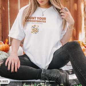 White Comfort Colors t-shirt printed with the words Count Your Carbohydrates Not Your Blessings and a drawing of a golden-brown braided pretzel with coarse salt grains, worn by a woman
