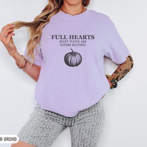 Orchid Comfort Colors t-shirt printed with the words Full Hearts Heavy Plates And Second Helpings and a drawing of a pumpkin surrounded by fall leaves and small acorns, worn by a woman