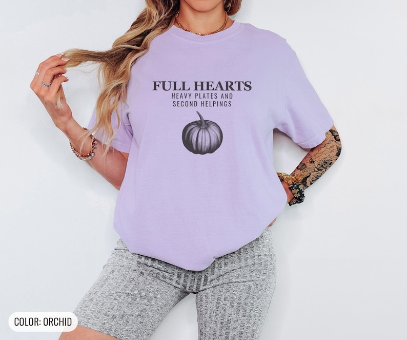 Orchid Comfort Colors t-shirt printed with the words Full Hearts Heavy Plates And Second Helpings and a drawing of a pumpkin surrounded by fall leaves and small acorns, worn by a woman