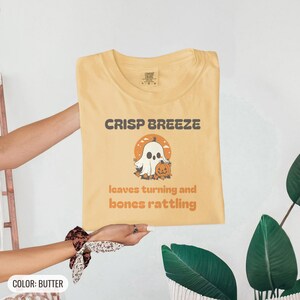 Butter Comfort Colors t-shirt printed with the words Crisp Breeze Leaves Turning And Bones Rattling and a drawing of a large cute smiling ghost beside a carved jack-o-lantern, folded flat lay