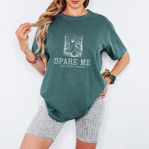 Blue Spruce Comfort Colors t-shirt printed with the words Spare Me Your Ghost Stories and a drawing of a friendly sheet ghost rolling a bowling ball towards a set of bowling pins, worn by a woman
