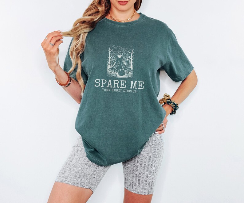 Blue Spruce Comfort Colors t-shirt printed with the words Spare Me Your Ghost Stories and a drawing of a friendly sheet ghost rolling a bowling ball towards a set of bowling pins, worn by a woman