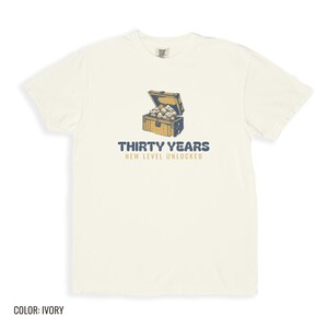 Ivory Comfort Colors t-shirt printed with the words Thirty Years New Level Unlocked and a drawing of a pixel-art treasure chest bursting open with light, folded flat lay