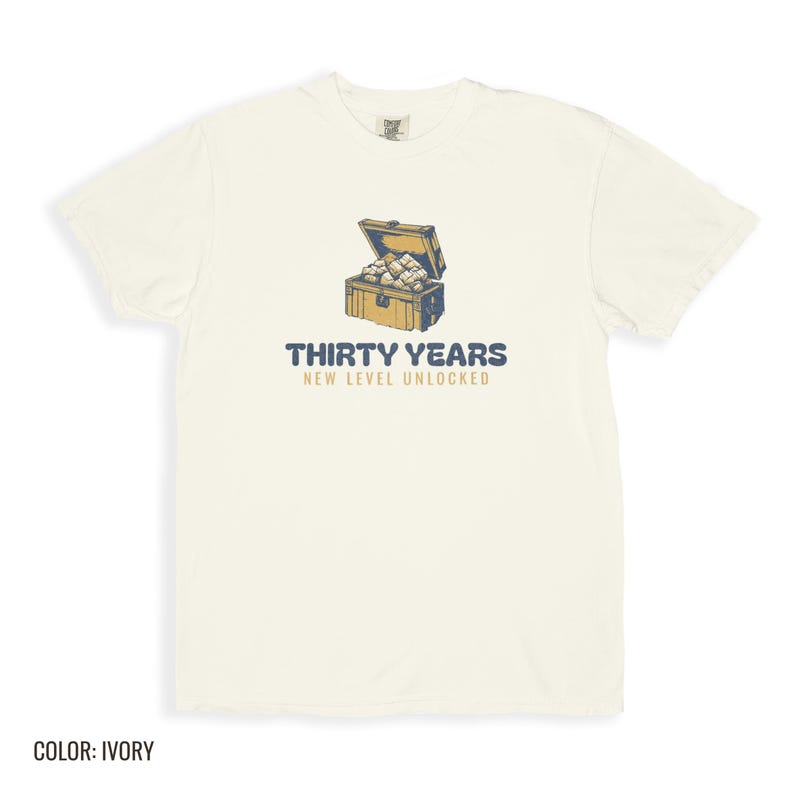 Ivory Comfort Colors t-shirt printed with the words Thirty Years New Level Unlocked and a drawing of a pixel-art treasure chest bursting open with light, folded flat lay