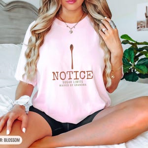 Blossom Comfort Colors t-shirt printed with the words Notice Sugar Limits Waived By Grandma and a drawing of a painted wooden spoon crossed over a cinnamon stick bundle, worn by a woman