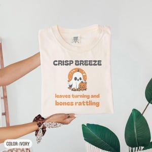 Ivory Comfort Colors t-shirt printed with the words Crisp Breeze Leaves Turning And Bones Rattling and a drawing of a large cute smiling ghost beside a carved jack-o-lantern, folded flat lay