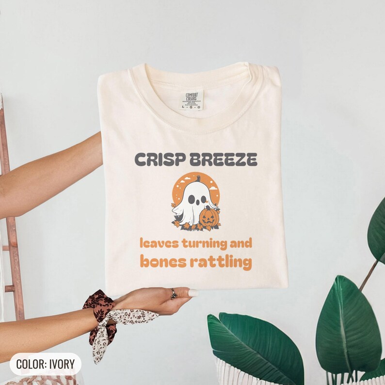 Ivory Comfort Colors t-shirt printed with the words Crisp Breeze Leaves Turning And Bones Rattling and a drawing of a large cute smiling ghost beside a carved jack-o-lantern, folded flat lay