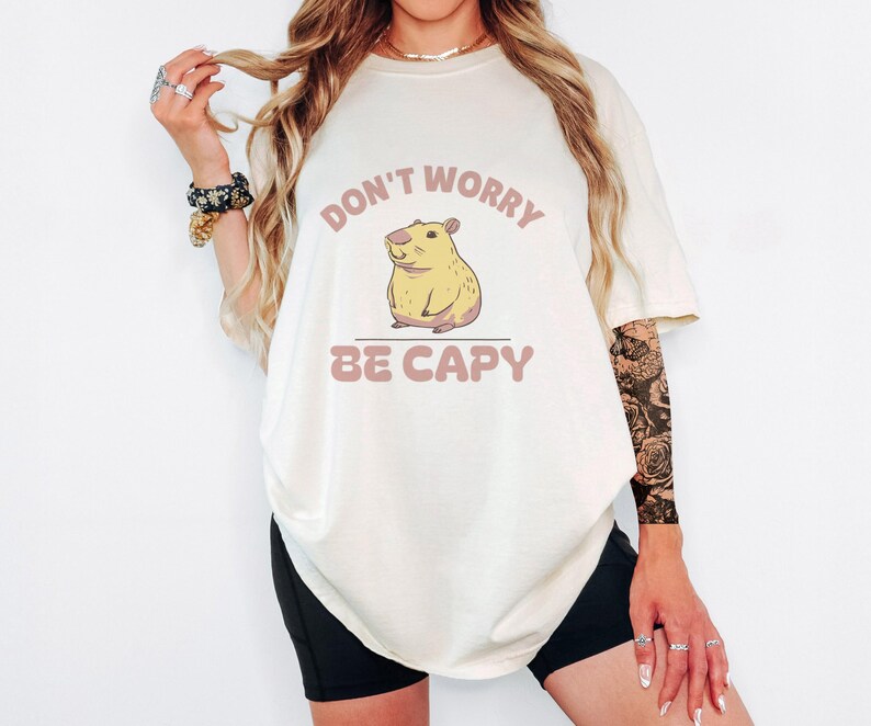 Ivory Comfort Colors t-shirt printed with the words Don'T Worry Be Capy and a drawing of a happy capybara with a small orange citrus slice on its head beside a green lily pad, worn by a woman