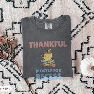 Pepper Comfort Colors t-shirt printed with the words Thankful Mostly For Recess and a drawing of a stack of retro school books topped with a glossy red apple and a steaming ceramic coffee, worn by a woman
