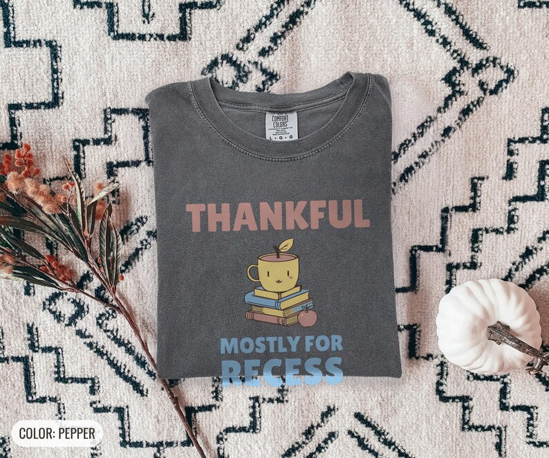 Pepper Comfort Colors t-shirt printed with the words Thankful Mostly For Recess and a drawing of a stack of retro school books topped with a glossy red apple and a steaming ceramic coffee, worn by a woman