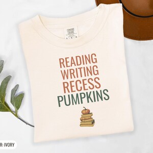 Ivory Comfort Colors t-shirt printed with the words Reading Writing Recess Pumpkins and a drawing of a stack of three vintage schoolbooks with a small orange pumpkin sitting on top, folded flat lay