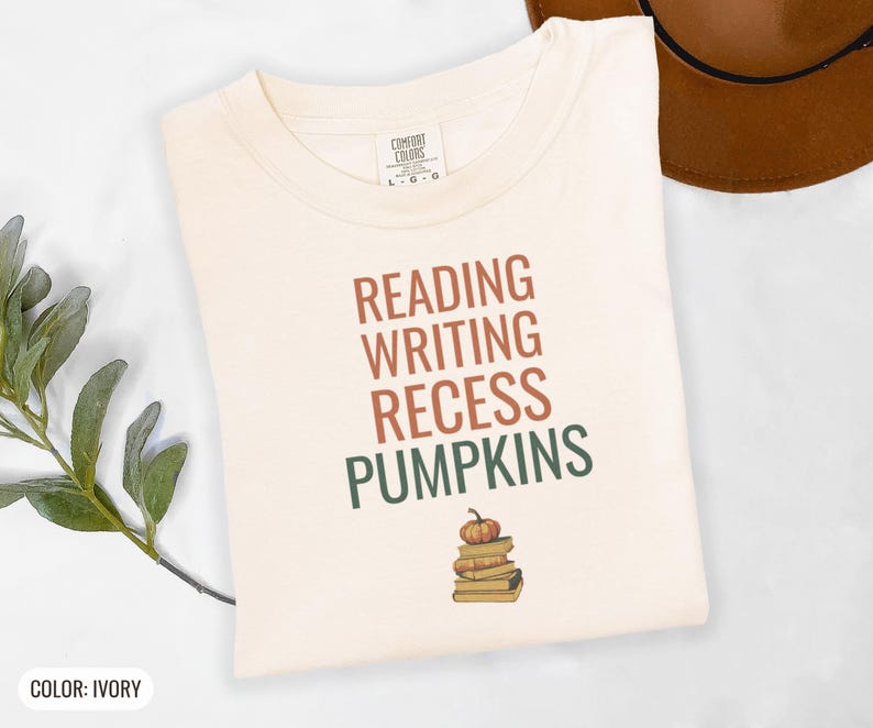 Ivory Comfort Colors t-shirt printed with the words Reading Writing Recess Pumpkins and a drawing of a stack of three vintage schoolbooks with a small orange pumpkin sitting on top, folded flat lay