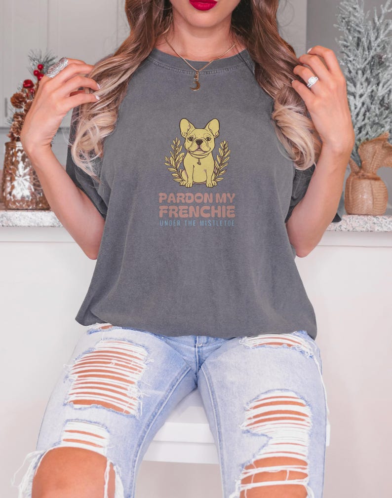 Pepper Comfort Colors t-shirt printed with the words Pardon My Frenchie Under The Mistletoe and a drawing of a vintage brass French Bulldog bottle opener lying next to a sprig of red-berried mistleto, worn by a woman