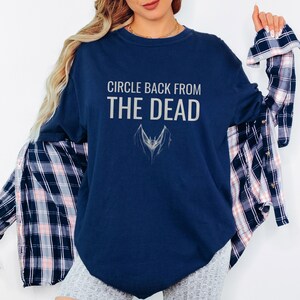 True Navy Comfort Colors t-shirt printed with the words Circle Back From The Dead and a drawing of a vintage silver paperclip bent into the shape of a bat, worn by a woman